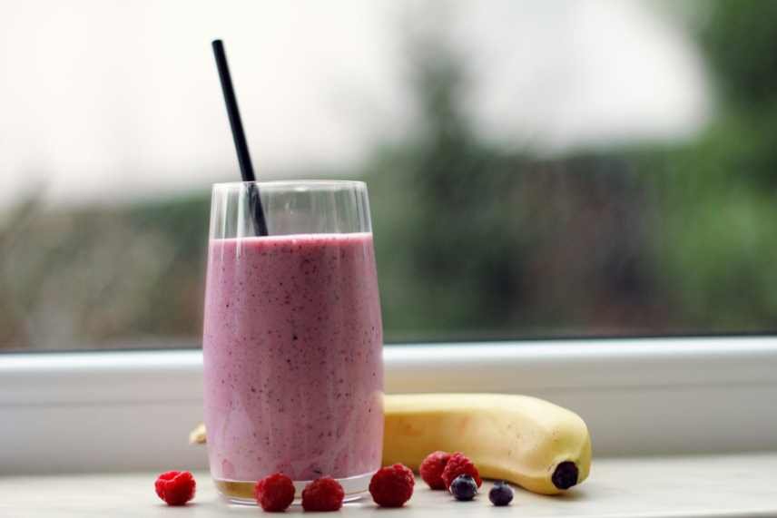 A mixed fruit smoothie on a window ledge with raspberries and a banana.