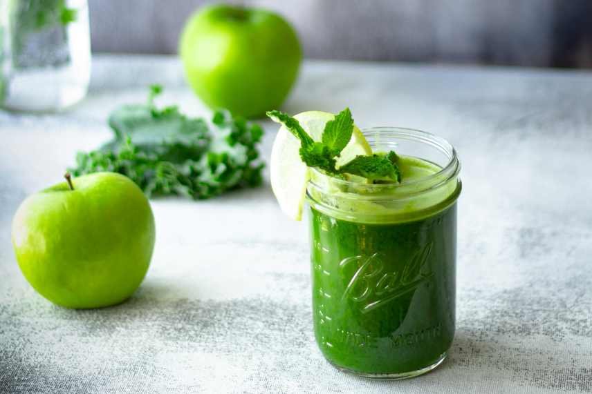 A jar containing a Kale and Apple smoothie, with kale and apples on the table.