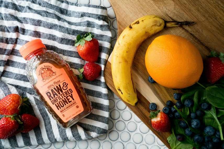 A bottle of Raw unfiltered honey, strawberries, a banana, an orange and blueberries on a table with a black and white cloth beneath some of them.