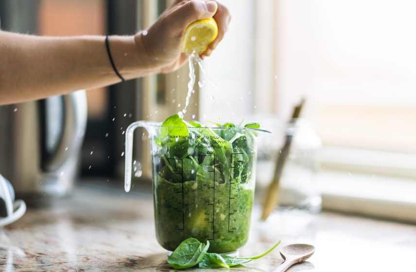A lemon being squeezed on top of spinach in a blender jug, ready to be blended into a green smoothie.