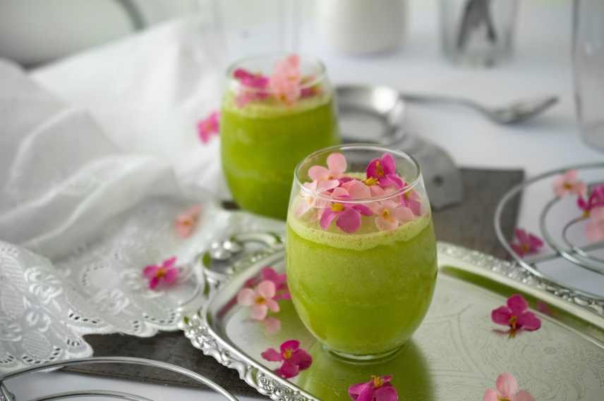 A green smoothie sits on a silver tray with pink flower petals sprinkled about.