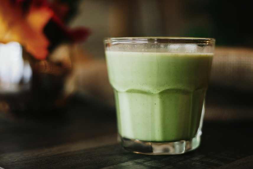 A close up of a green smoothie in a small glass on a dark table. The background is blurred out.