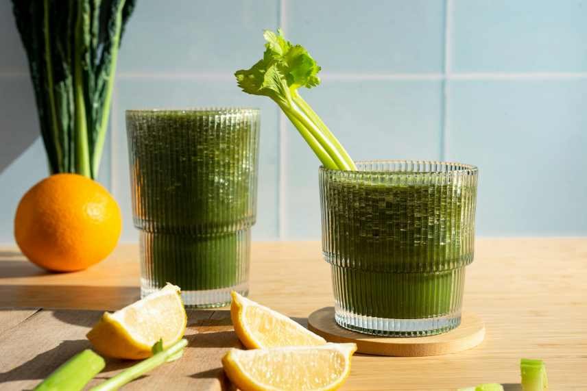 Two green smoothies with a stick of celery, kale, an orange and lemon wedges, on a wooden table with a light blue tiled background.