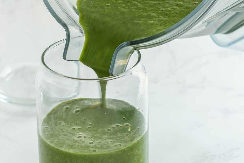 Close up photo of a creamy avocado and spinach smoothie being poured into a glass on  white bench.