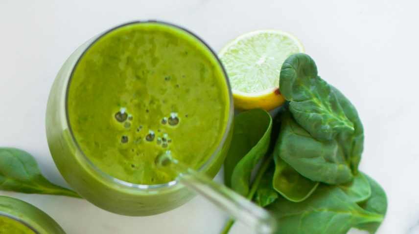 A Mango and Spinach smoothie in a tall glass with a glass straw. There are some spinach leaves and a lime sitting on the table at the base of the glass.