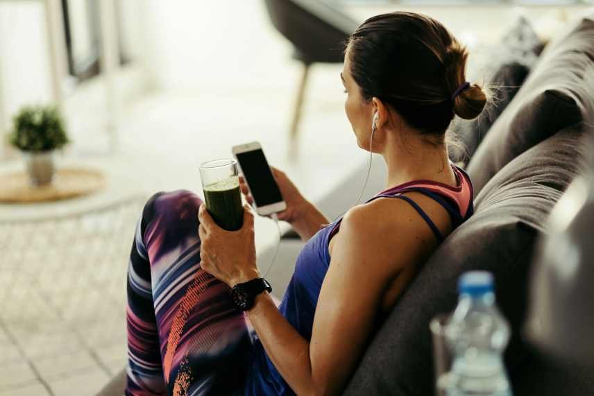 A female in fitness clothing sitting on a couch listening to a podcast while holding a healthy smoothie for energy and weight loss.