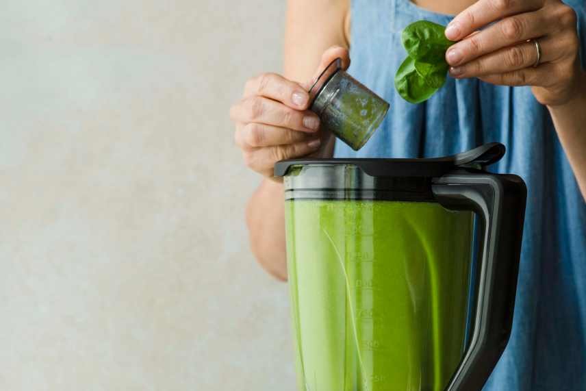 The top section of a blender full of a heart healthy green smoothie mix. A female is adding more spinach via the lid opening.