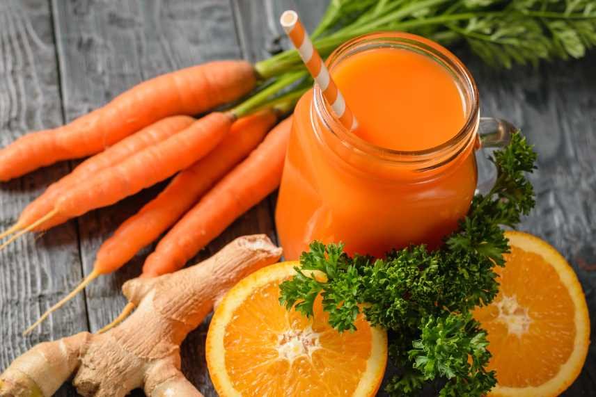 A mason jar of fresh carrot smoothie with a cocktail straw, parsley, ginger root, orange halves and a bunch of carrots.